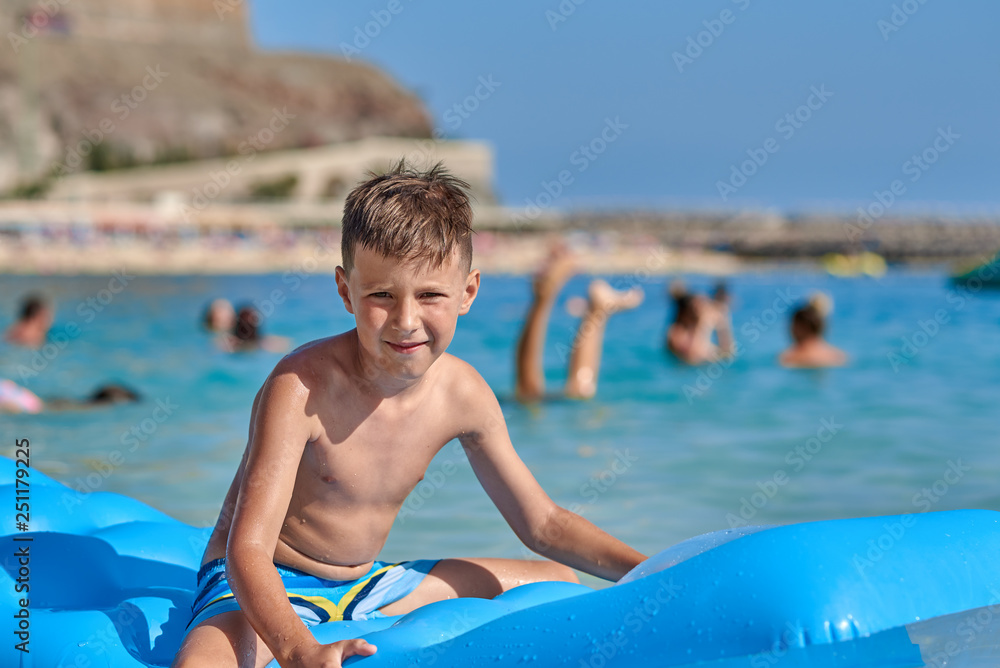 Good-looking European boy is sitting on the inflatable matress in the sea and smiling to the camera.