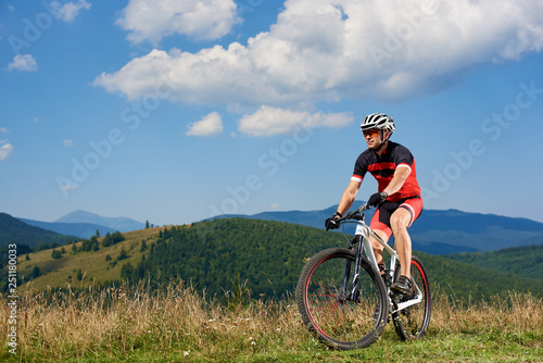 Professional sportsman cyclist in sportswear and helmet cycling a bike in high grass, enjoying beautiful view of Carpathian mountains on sunny summer day. Active lifestyle and extreme sport concept