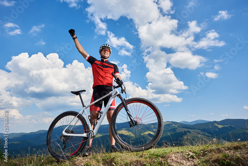 Happy professional sportsman cyclist standing with cross country bicycle on a hill, rasing hand, against blue sky with clouds on summer sunny day. Outdoor sport, success and life goals concept