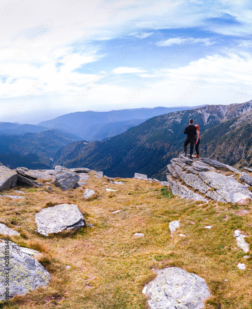 Back view of tourist couple, man and girl stand on rocky mountain top enjoying breathtaking autumn mountain panorama. Tourism, traveling lifestyle concept, vertical photo, Romania Transalpina