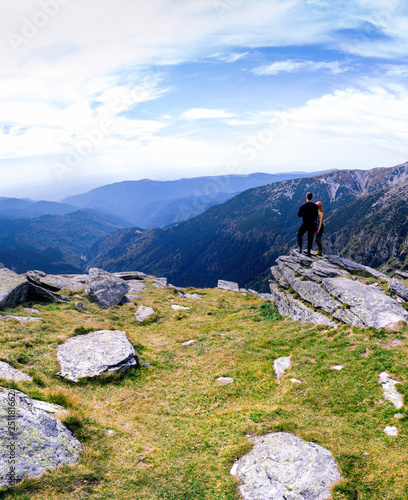 Back view of tourist couple, man and girl stand on rocky mountain top enjoying breathtaking summer mountain panorama. Tourism, traveling lifestyle concept. motorcyclists, vertical photo, Romania