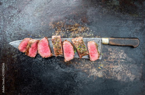Traditional barbecue aged venison backstrap roast sliced with herbs as top view on a large knife with rustic background with copy space photo