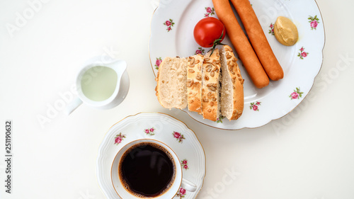 Parówki, pokrojona bułka ze słonecznikiem, pomidor, kawa leżące na białym tle