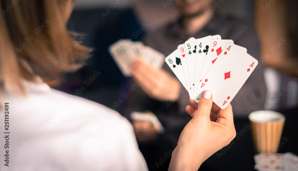 Friends are playing cards together at home. Woman is holding cards in her hands, man in the blurry background. Friends are playing cards together at home. Woman is holding cards in her hands, man in the blurry background.