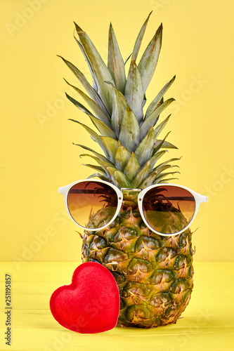 Pineapple in sunglasses and red heart. Fresh pineapple on yellow background.