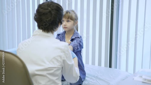 Little girl visiting doctor for medical checkup: female pediatrician in white coat and gloves rolling up her sleeves and palpating her arm