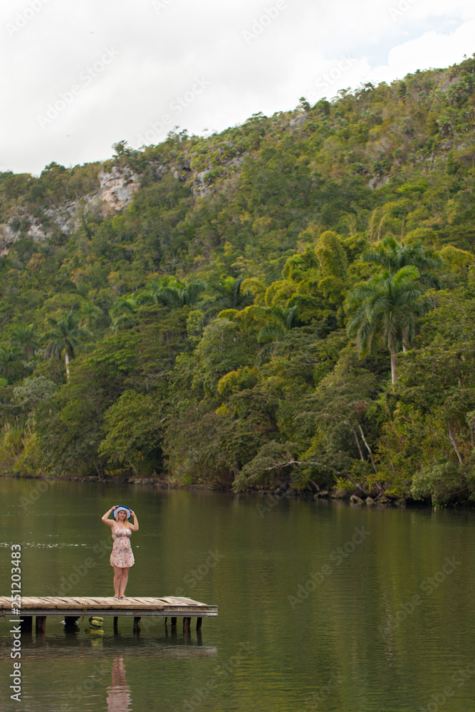Fototapeta premium Young blonde woman in summer dress at pier near wild river in jungle