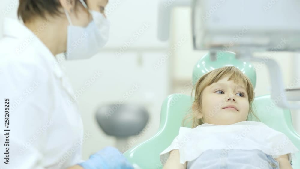 Child patient lying in dental chair and opening mouth preparing to be examined by female doctor in white coat and medical mask Child patient lying in dental chair and opening mouth preparing to be examined by female doctor in white coat and medical mask