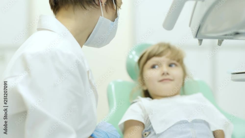 Anonymous female dentist in mask explaining treatment procedure to adorable little girl in doctor office Anonymous female dentist in mask explaining treatment procedure to adorable little girl in doctor office
