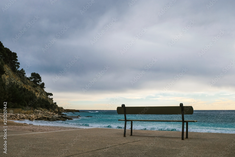 Silhouette of an empty wooden bench on the coast of the Mediterranean Sea in winter under gray clouds. Solitude. Pandemic concept. Spreading concept.