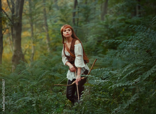 gorgeous lady with long red hair in leather clothes follows wild animal, hunts down prey in rainforest, rite of initiation into hunters, girl pulled bowstring of bow with arrow, ready to shoot photo