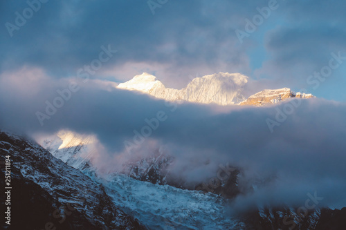 Sunlight wins over clouds above the Himalayan Peaks, Annapurna Circuit Trek, Nepal. Slopes covered with mist. Sharp slopes. Smaller mountain in front, covered in shadow. Overcast but sunny.