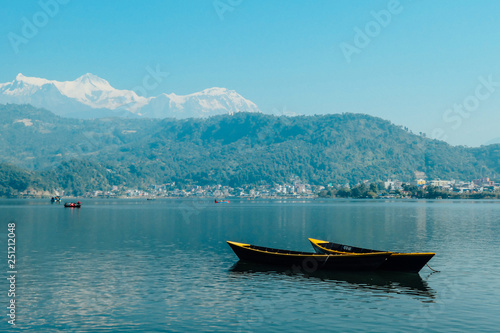 Two empty boats floating on the surface of Phewa Lake, Pokhara, Nepal. IN the back more boats with tourists. Cityscape on the shore. High Himalayan peaks in the back. Chilled and relaxed atmosphere.