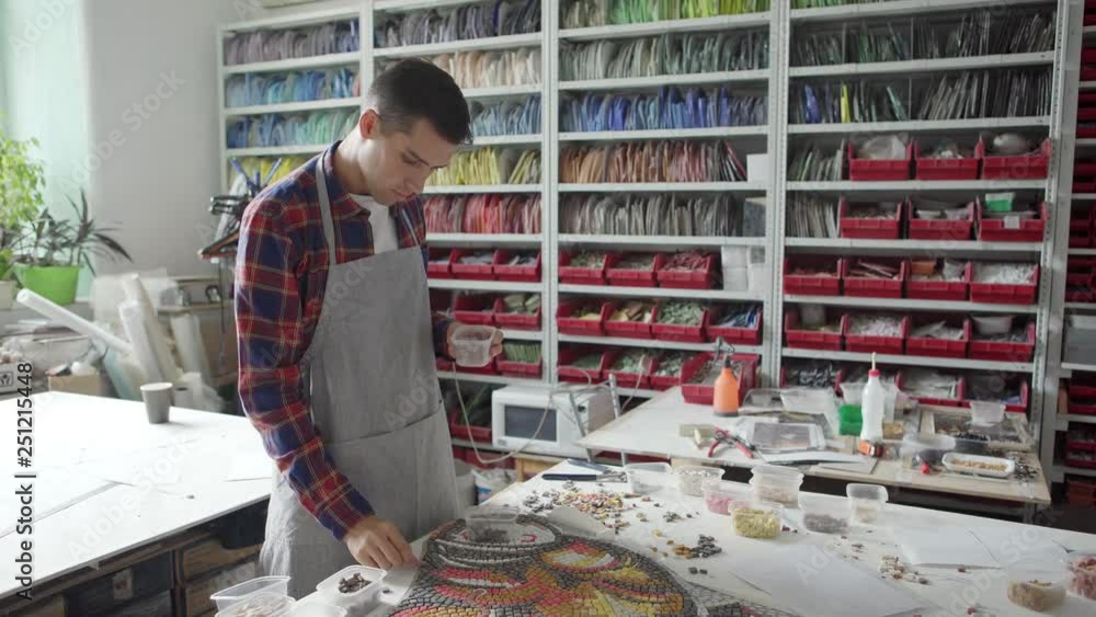 Young artisan in apron taking piece of mosaic from small box and putting in on table in workshop Young artisan in apron taking piece of mosaic from small box and putting in on table in workshop