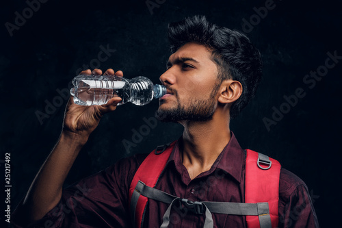 Handsome Indian hiker with backpack exhausted with thirst drinking water. Studio photo against a dark textured wall