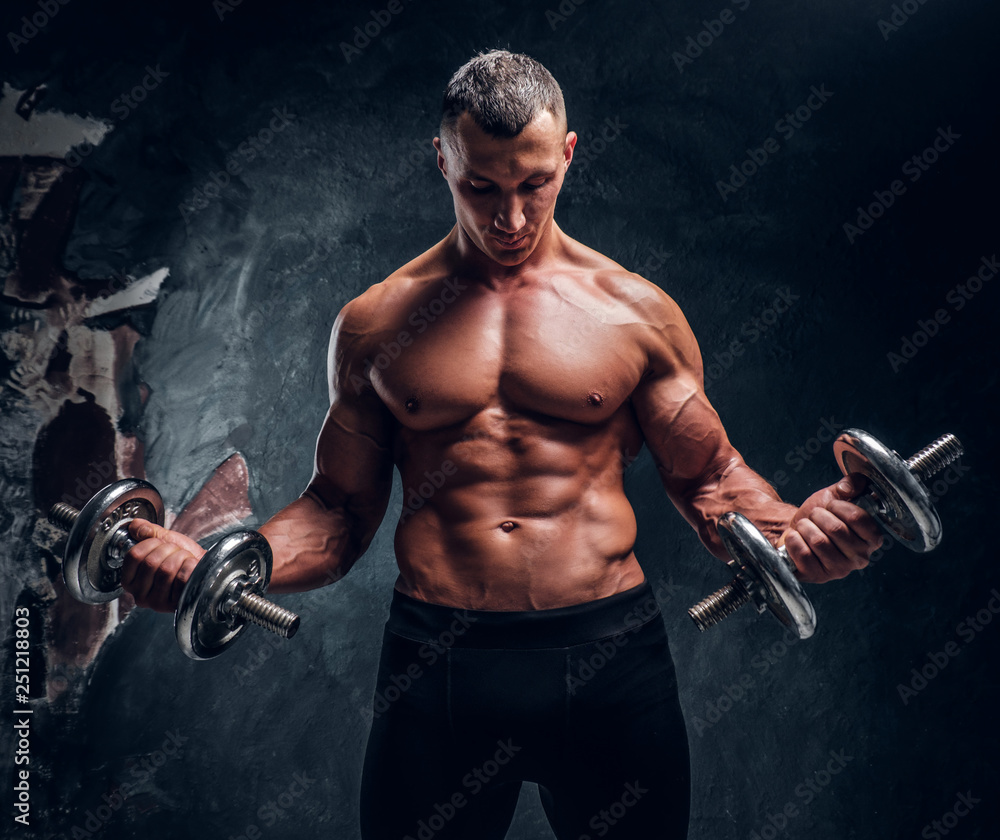 Muscular young guy trains with two dumbbells.  Studio photo with dark wall background