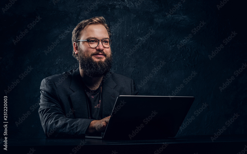 Smiling bearded programmer sitting at the table with laptop in office against a dark wall Smiling bearded programmer sitting at the table with laptop in office against a dark wall