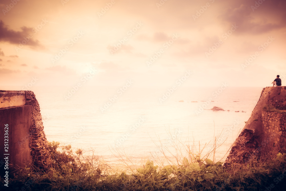 Lone traveler man on cliff looking with inspiration at horizon with sunlight during sunset with effect of light at the end of tunnel