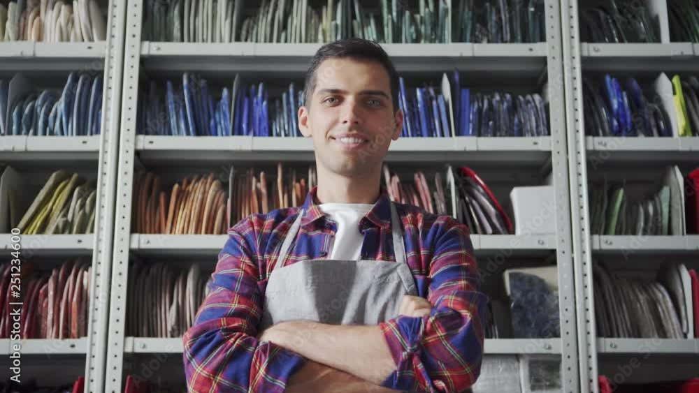 Attractive male in apron turning to camera and smiling after searching for mosaic samples on shelves in workshop Attractive male in apron turning to camera and smiling after searching for mosaic samples on shelves in workshop
