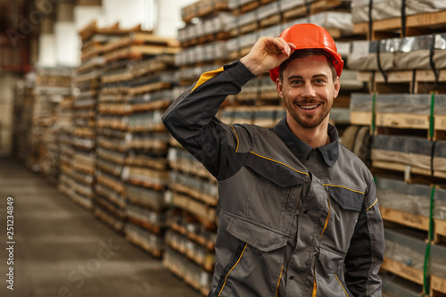 Fototapeta Cheerful handsome male engineer smiling to the camera while putting on protective hardhat, posing at the storage of metalworking factory