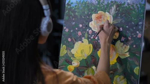 Close up back view of creative female artist in headphones listening to music and painting artwork of nature on easel. Large yellow peonies on canvas against green background photo