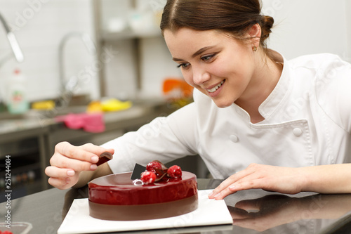 Beautiful and cute girl smiling and decorating glazed cake. Young confectioner wearing white uniform while working in confectionery. She baking delicious dark red with chocolate. photo