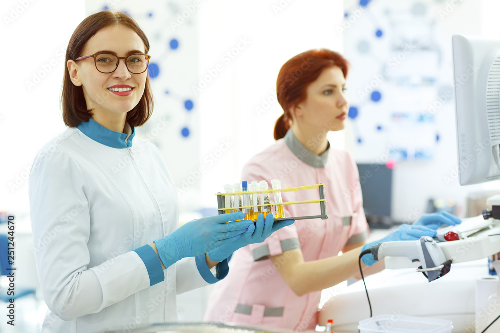 Beautiful female lab worker looking at camera and smiling. Woman in white uniform, blue gloves and spectacles holding test tubes. Other lab technician working with computer on background. Beautiful female lab worker looking at camera and smiling. Woman in white uniform, blue gloves and spectacles holding test tubes. Other lab technician working with computer on background.