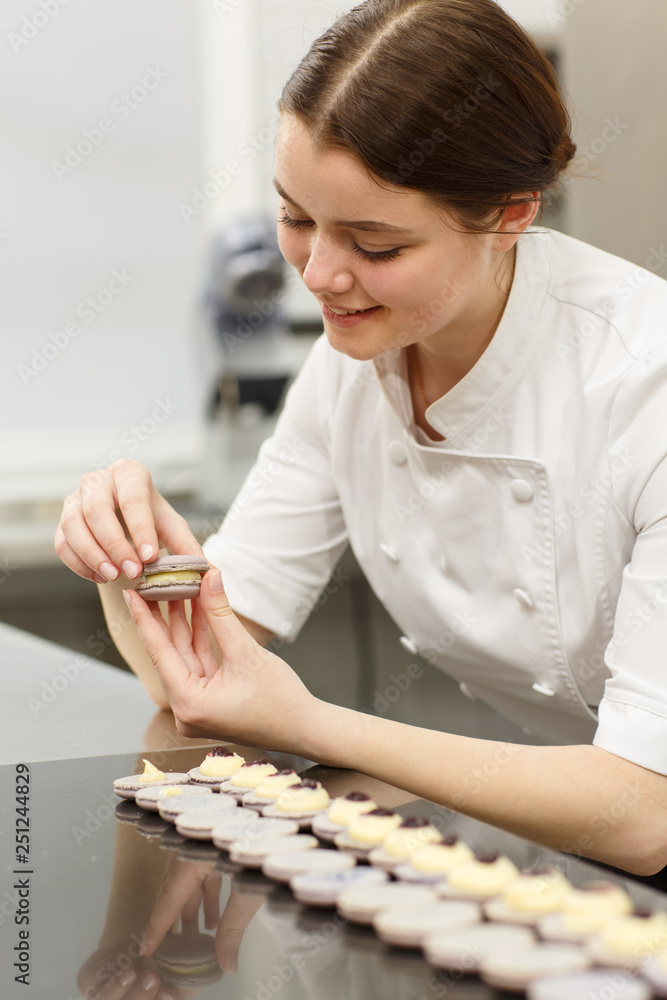 Cute and beautiful young girl wearing white uniform and working in confectionery. Female confectioner joining two parts of cookies and making macarons. Row of delicious macarons placed on table. Cute and beautiful young girl wearing white uniform and working in confectionery. Female confectioner joining two parts of cookies and making macarons. Row of delicious macarons placed on table.
