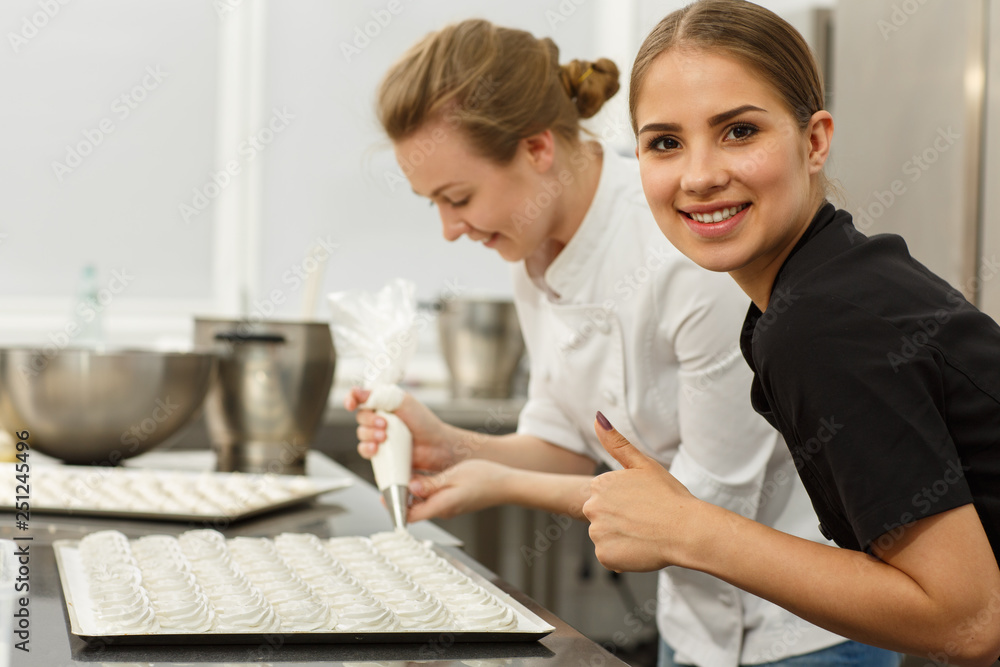 Fototapeta premium Beautiful women wearing white uniform, holding confectionery bags and putting dough on baking sheet, other gesturing super by big finger. Two confectioners cooking desserts.