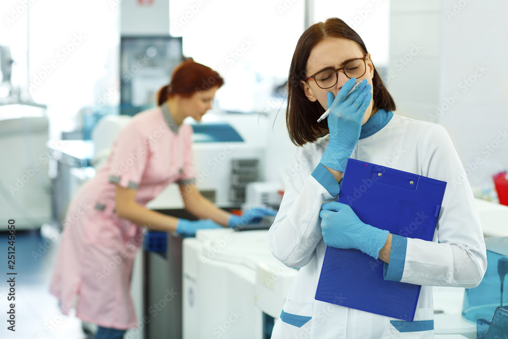Female lab technician yawning while working at modern research laboratory. Woman in blue gloves and spectacles holding dark blue folder. Concept of medical and pharmacology industry. Female lab technician yawning while working at modern research laboratory. Woman in blue gloves and spectacles holding dark blue folder. Concept of medical and pharmacology industry.