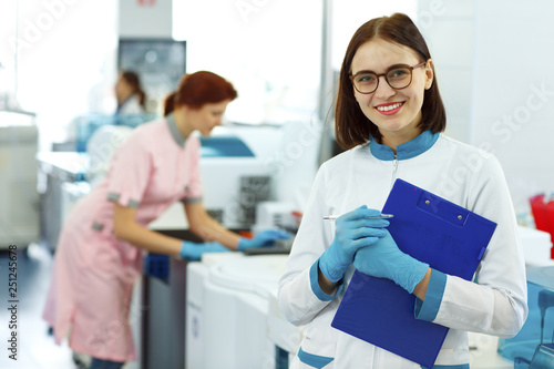 Front view of beautiful lab technician looking at camera and posing. Woman smiling  wearing white uniform  blue gloves and holding dark blue folder. Other medical staff working on background.