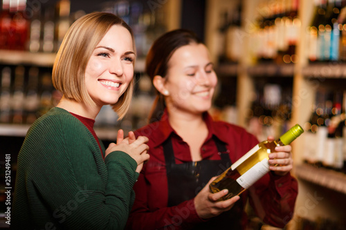 Image of two smiling women with bottle of wine in store on background of shelves