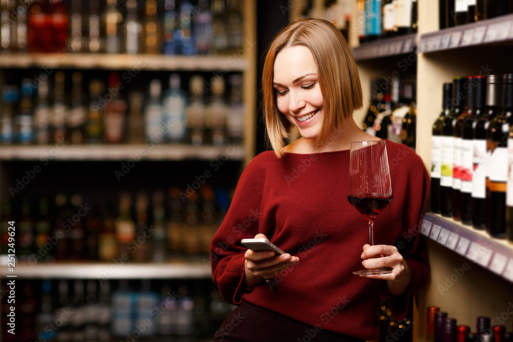 Image of blonde with phone and glass in hands at store with wine