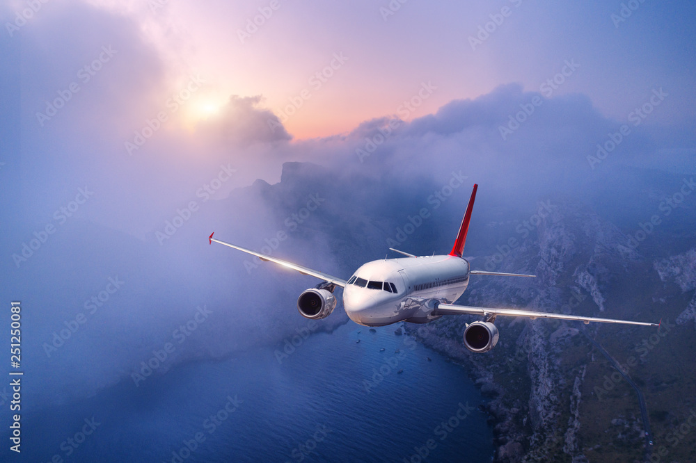 Passenger airplane is flying over clouds at sunset. Landscape with white airplane, low clouds, sea coast, purple sky at dusk. Aircraft is landing. Business trip. Commercial plane. Travel. Aerial view Passenger airplane is flying over clouds at sunset. Landscape with white airplane, low clouds, sea coast, purple sky at dusk. Aircraft is landing. Business trip. Commercial plane. Travel. Aerial view