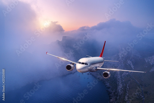 Passenger airplane is flying over clouds at sunset. Landscape with white airplane, low clouds, sea coast, purple sky at dusk. Aircraft is landing. Business trip. Commercial plane. Travel. Aerial view