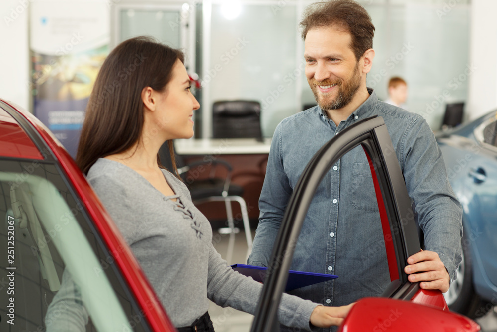 Positivity and friendly man manager showing young happy woman customer cars. Beautiful smiling girl sitting inside vehicle in red car. Bearded sales consultant working in car dealership. Positivity and friendly man manager showing young happy woman customer cars. Beautiful smiling girl sitting inside vehicle in red car. Bearded sales consultant working in car dealership.
