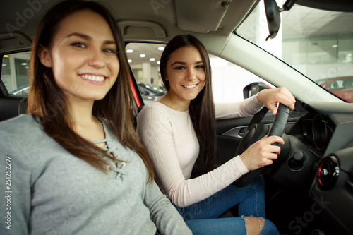 Gorgeous young girls sitting inside car, smiling and looking at camera. One of girls holding hands on steering wheel. Female customers of car dealership choosing auto in showroom. © Nestor