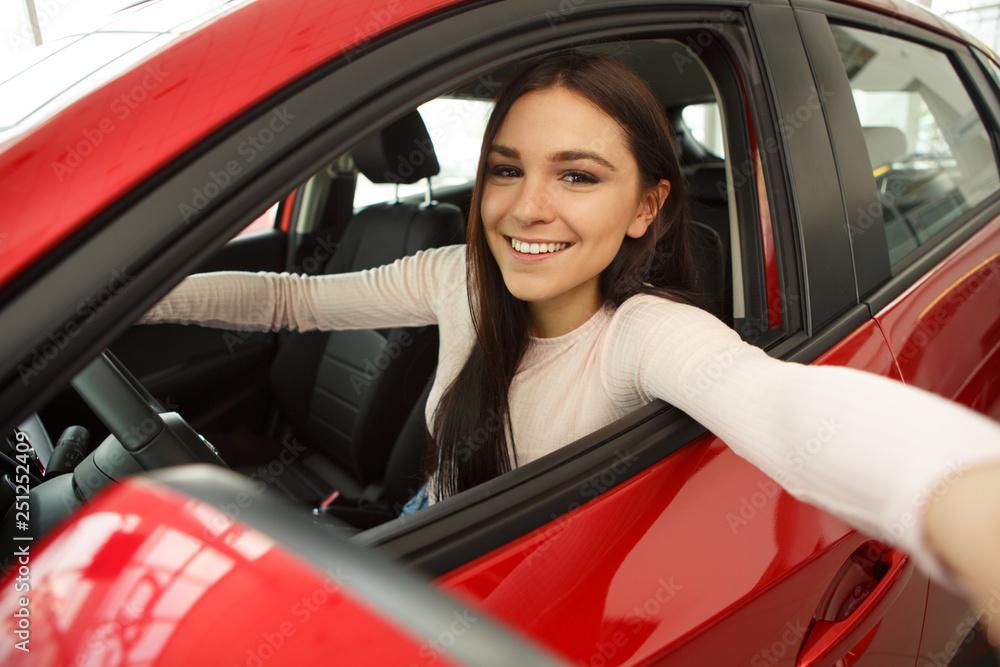 Happy young woman sitting in red car and making selfie. Beautiful girl with chestnut hair smiling and looking at camera. Pretty female customer making purchase of automobile.