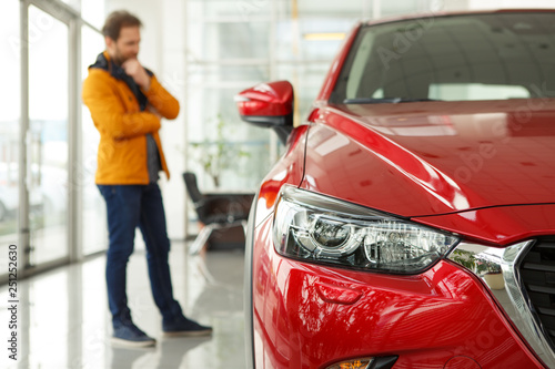 Front view of headlight of beautiful red automobile. Male customer choosing cars and thinking about features on background. Concept of car dealership, purchase and service.