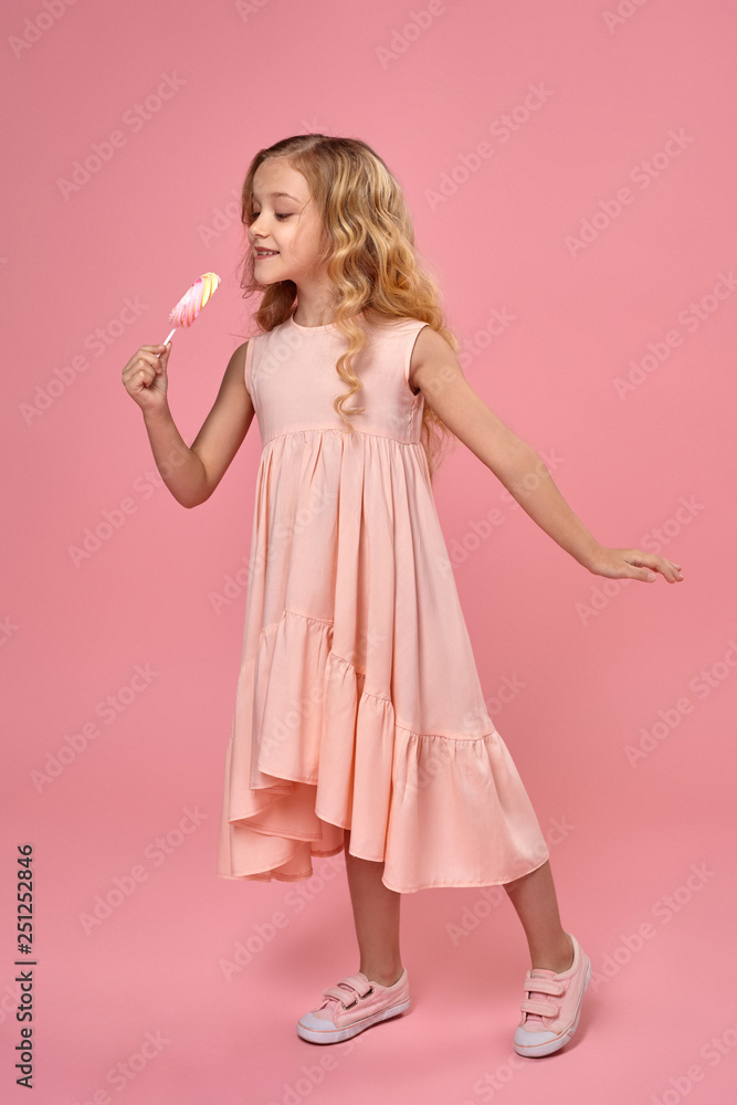 Little girl with a blond curly hair, in a pink dress is posing with a candy Little girl with a blond curly hair, in a pink dress is posing with a candy