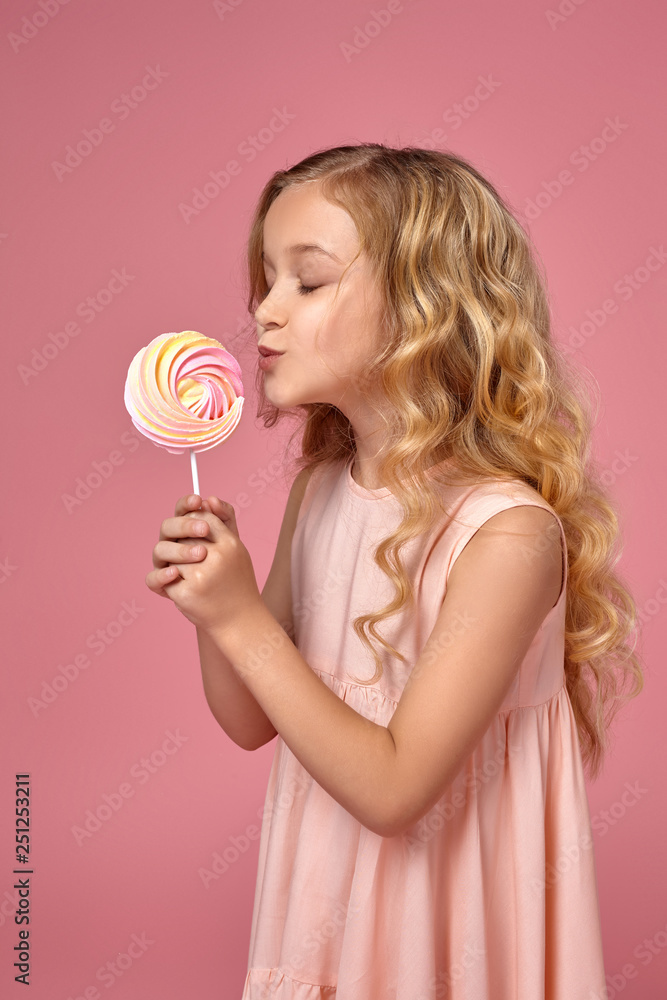 Little girl with a blond curly hair, in a pink dress is posing with a candy