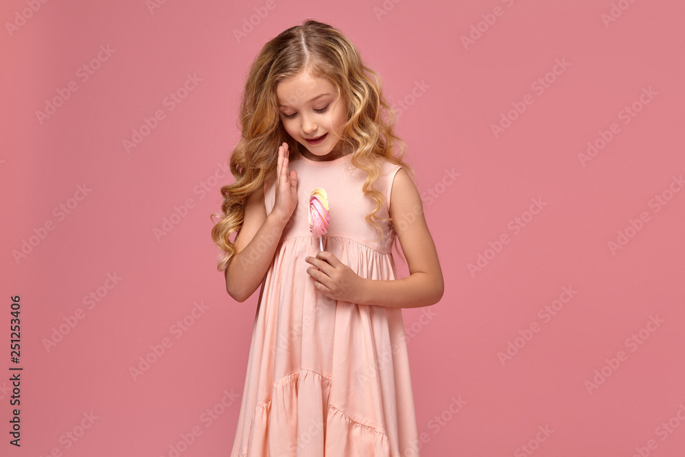 Little girl with a blond curly hair, in a pink dress is posing with a candy Little girl with a blond curly hair, in a pink dress is posing with a candy