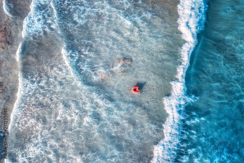 Aerial view of a young woman swimming with the donut swim ring in the clear blue sea with beautiful waves at sunset in summer. Tropical aerial landscape with girl, azure water. Top view. Travel © den-belitsky