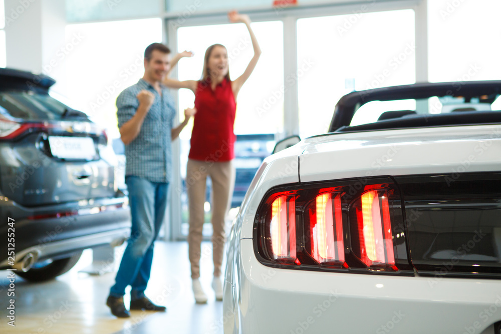Closeup of rear headlights of new expensive car in auto salon. Happy handsome male and redhaired beautiful woman standing near automobile, jumping and rejoicing in purchase on background.