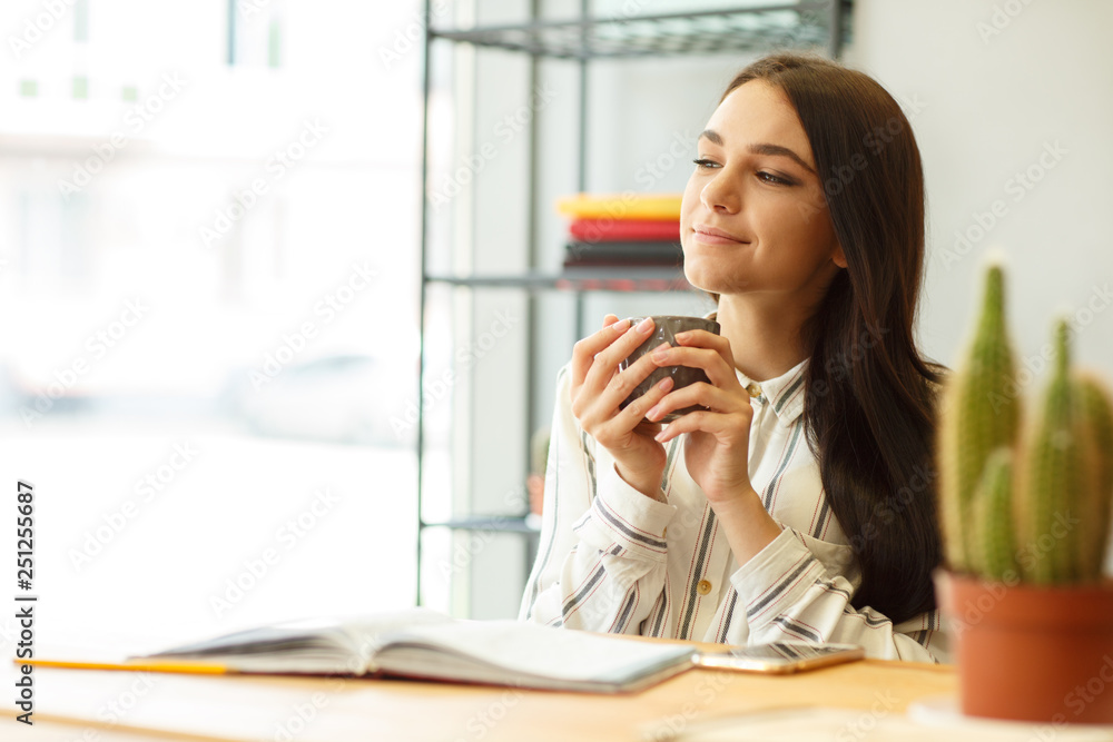 Portrait of dreamy designer with cup in hands sitting at desk in bright office. Portrait of dreamy designer with cup in hands sitting at desk in bright office.