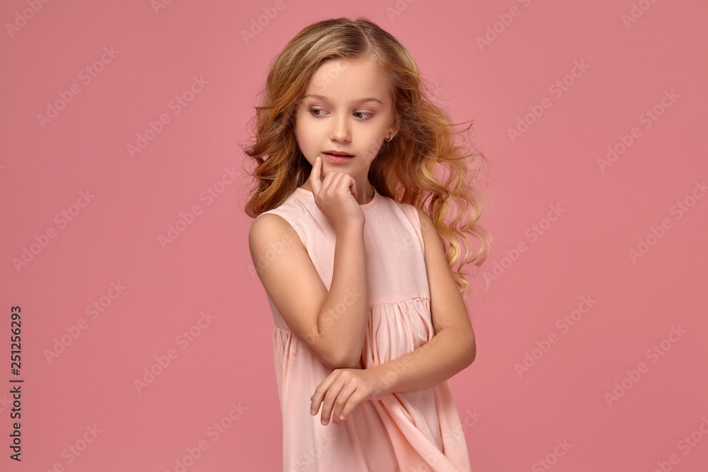 Little girl with a blond curly hair, in a pink dress is posing for the camera Little girl with a blond curly hair, in a pink dress is posing for the camera
