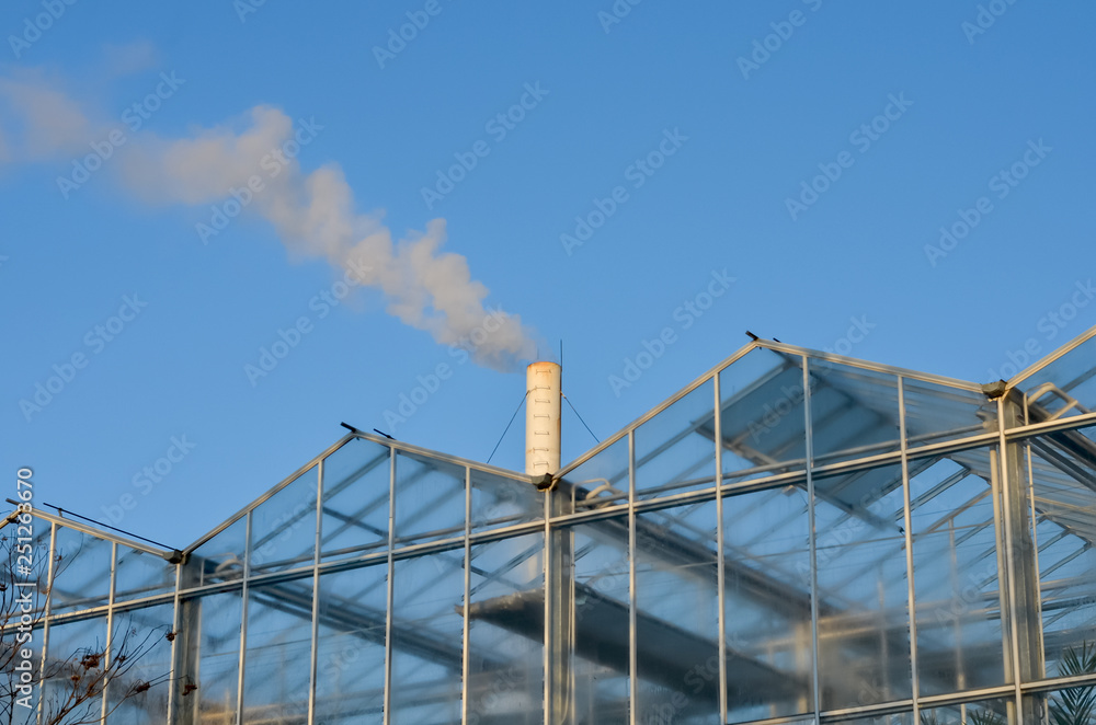 Glass  greenhouse with smoking  pipe emitting  smoke against blue sky . Heating the greenhouses in winter  .Environmental pollution.
