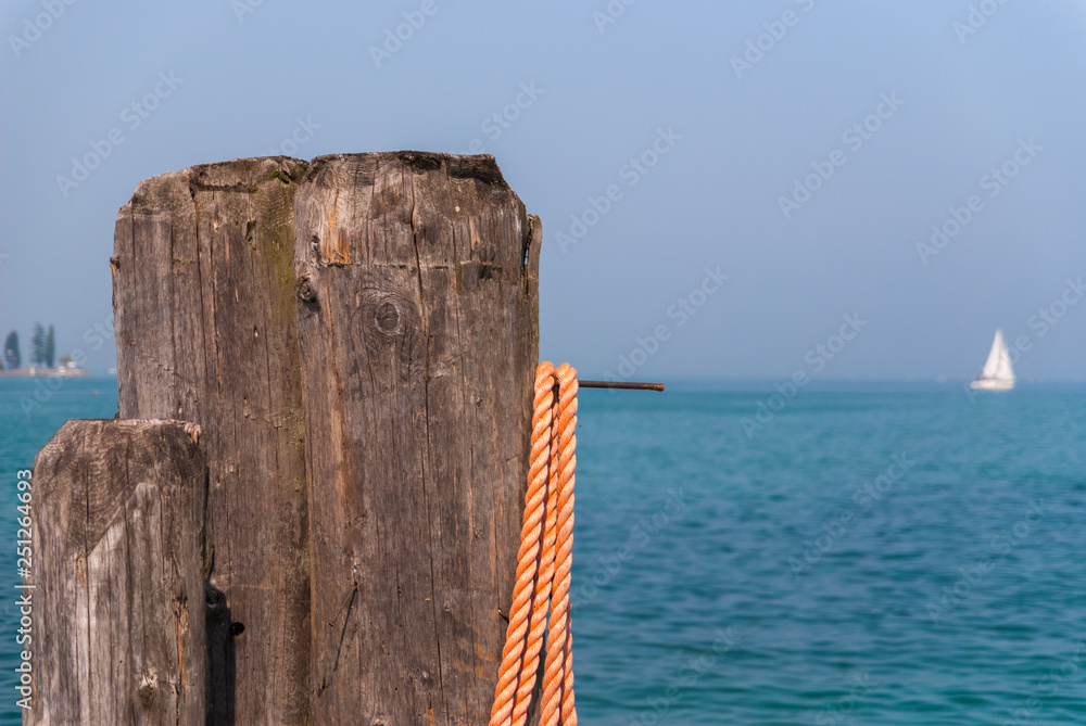 Wooden mooring pole with orange rope. In the background blurred Lake Garda with white sailboat