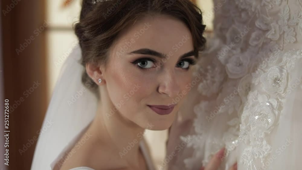 Beautiful and lovely bride in night gown stands near wedding dress and looks at camera. Close-up shot. Wedding morning. Pretty and well-groomed woman. Slow motion Beautiful and lovely bride in night gown stands near wedding dress and looks at camera. Close-up shot. Wedding morning. Pretty and well-groomed woman. Slow motion