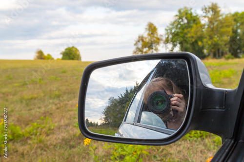 Car mirror, the reflection of the female photographer in it and landscape with field and tree